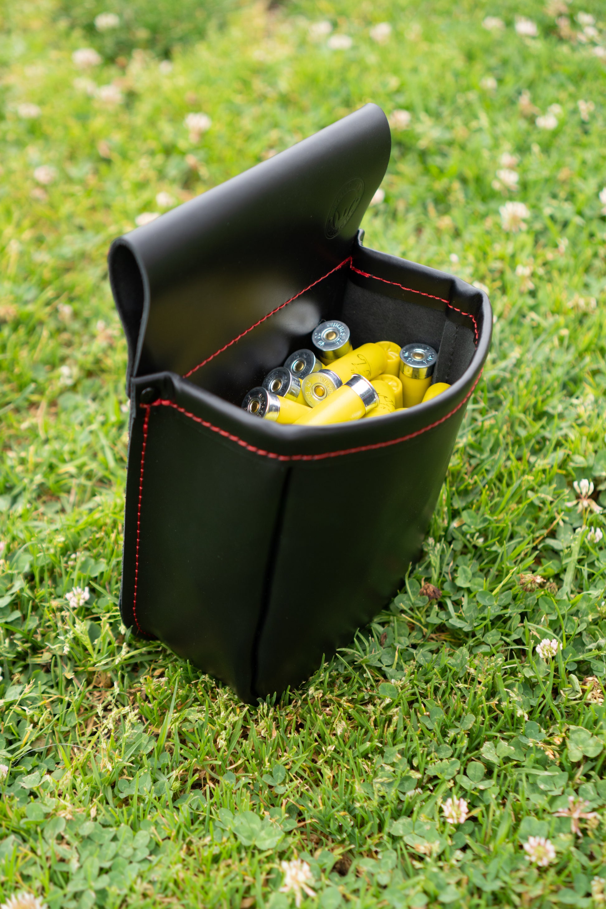 Black leather shotgun shell pouch with red stitching sitting upright in green grass