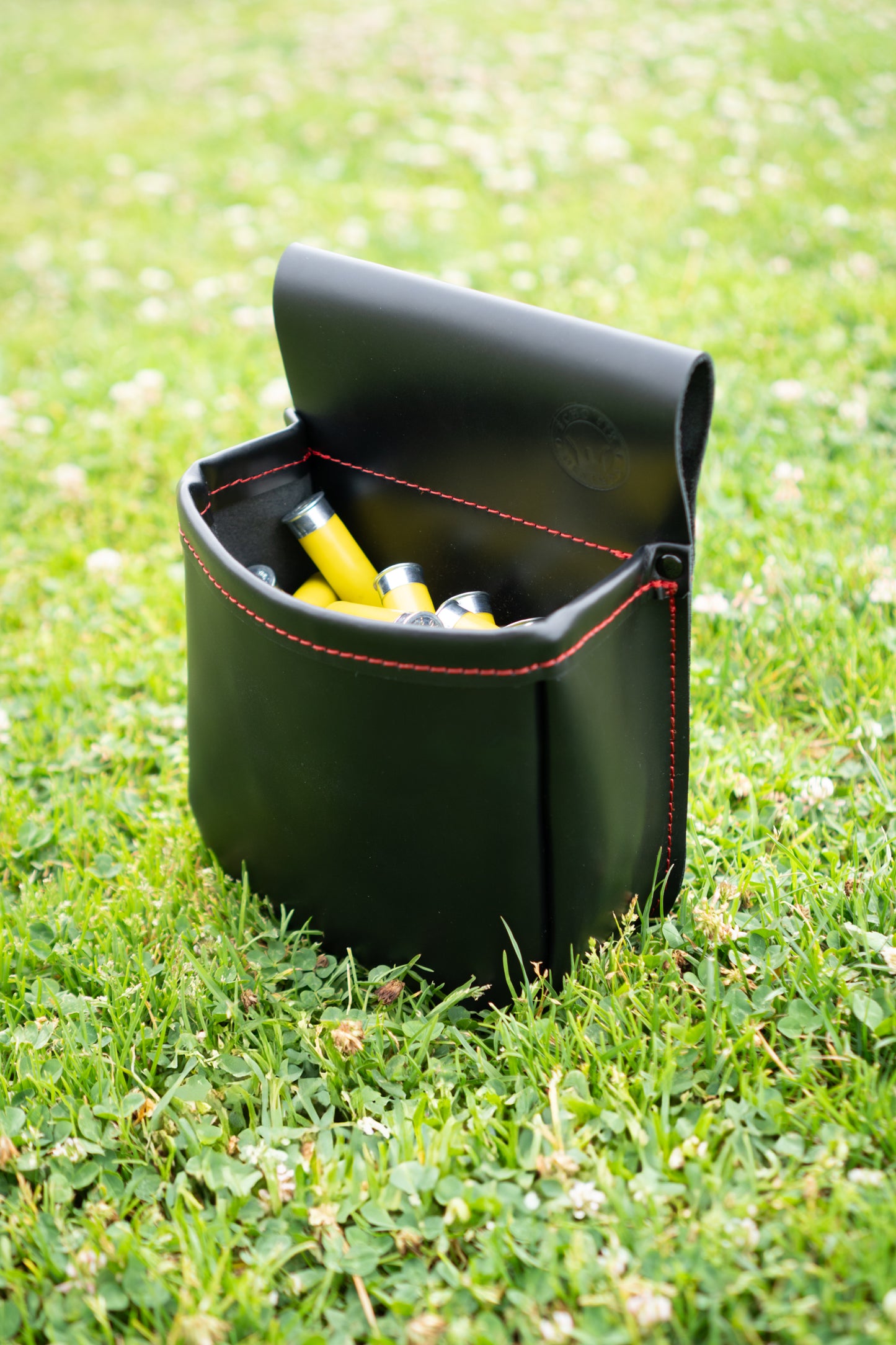 Black leather shotgun shell pouch with red stitching sitting upright in green grass