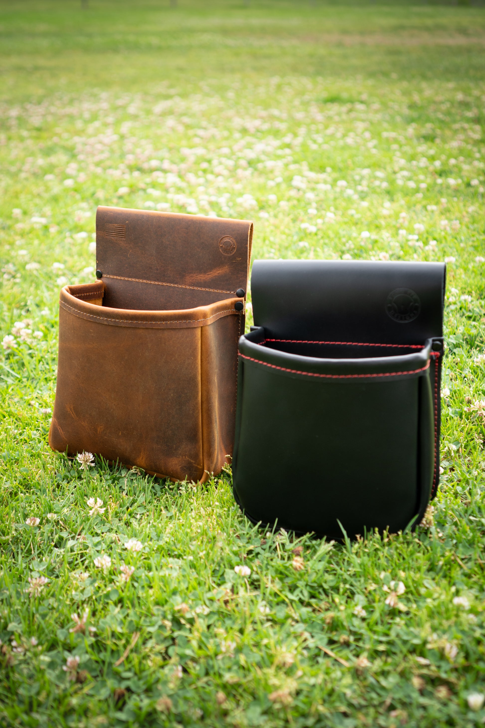 Black leather shotgun shell pouch with red stitching and brown leather shotgun shell pouch sitting upright in green grass