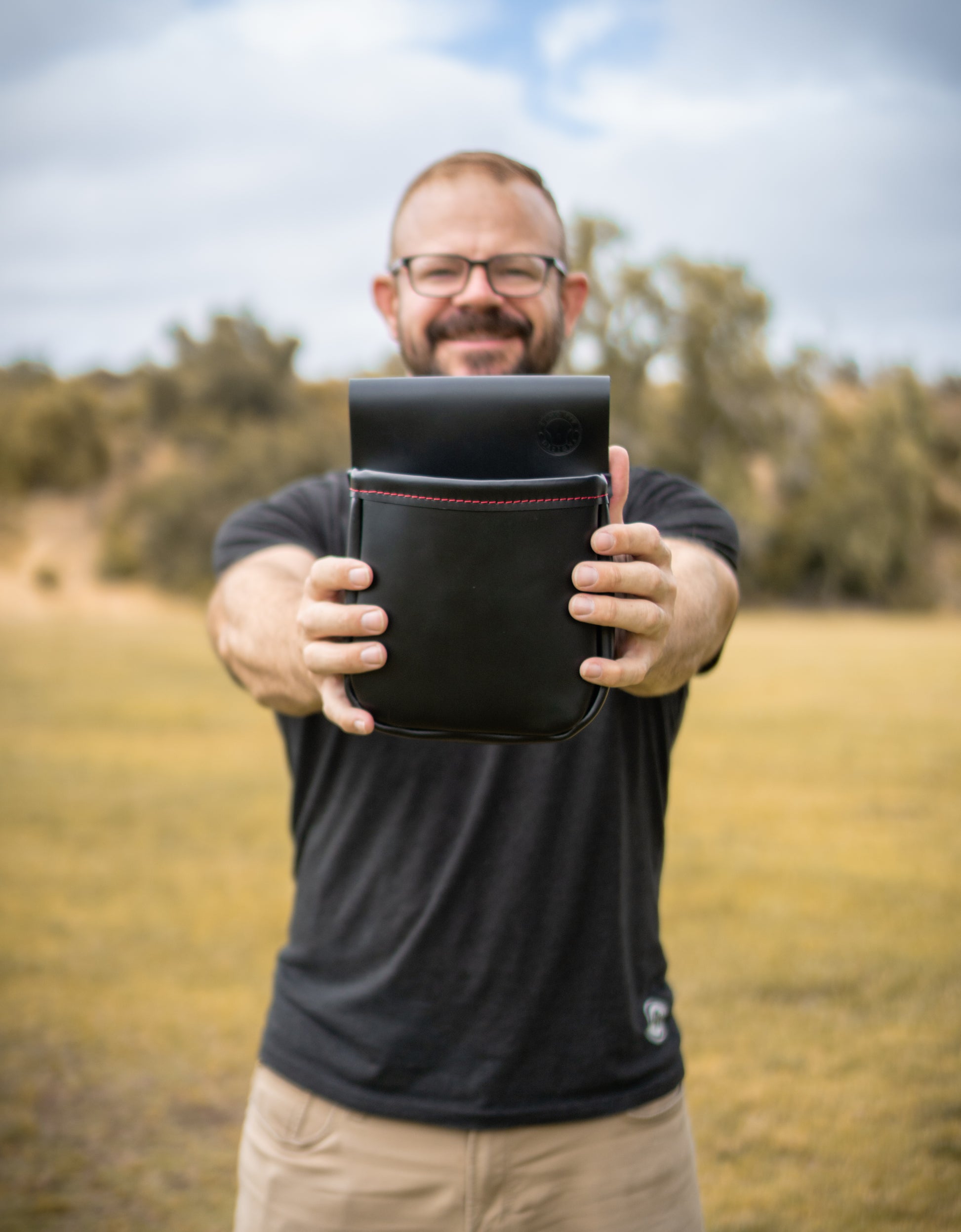 Man standing and holding out a black leather shotgun shell pouch with red stitching with both hands
