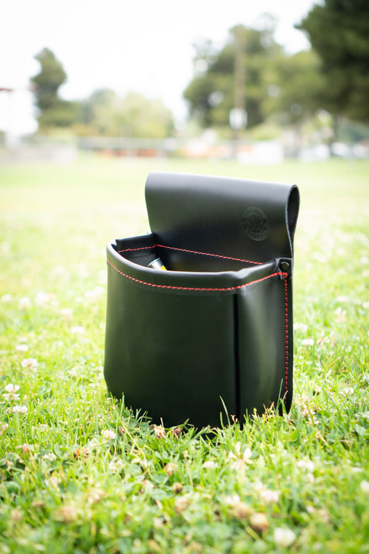 Black leather shotgun shell pouch with red stitching sitting upright in green grass