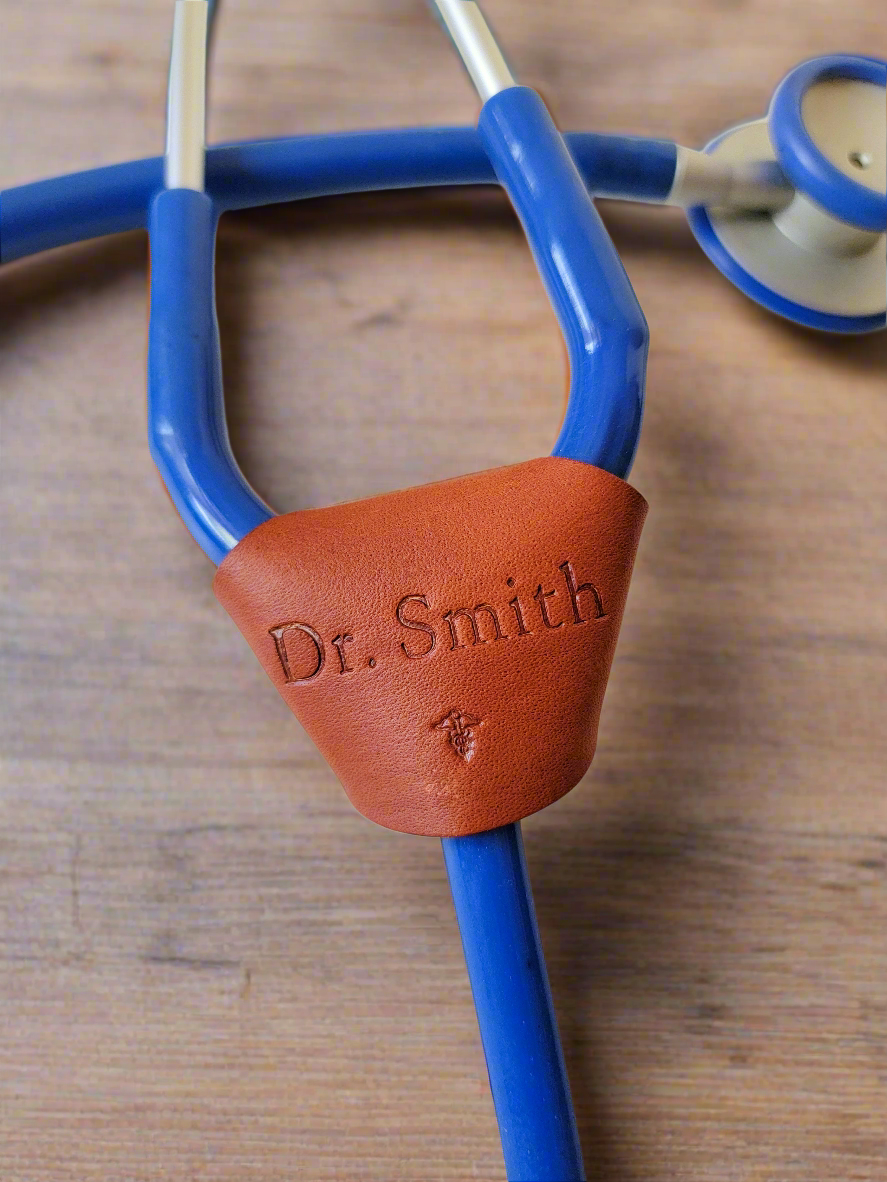 Handmade leather stethoscope tag in the colored tan, wrapped around a blue stethoscope laying on a wooden table